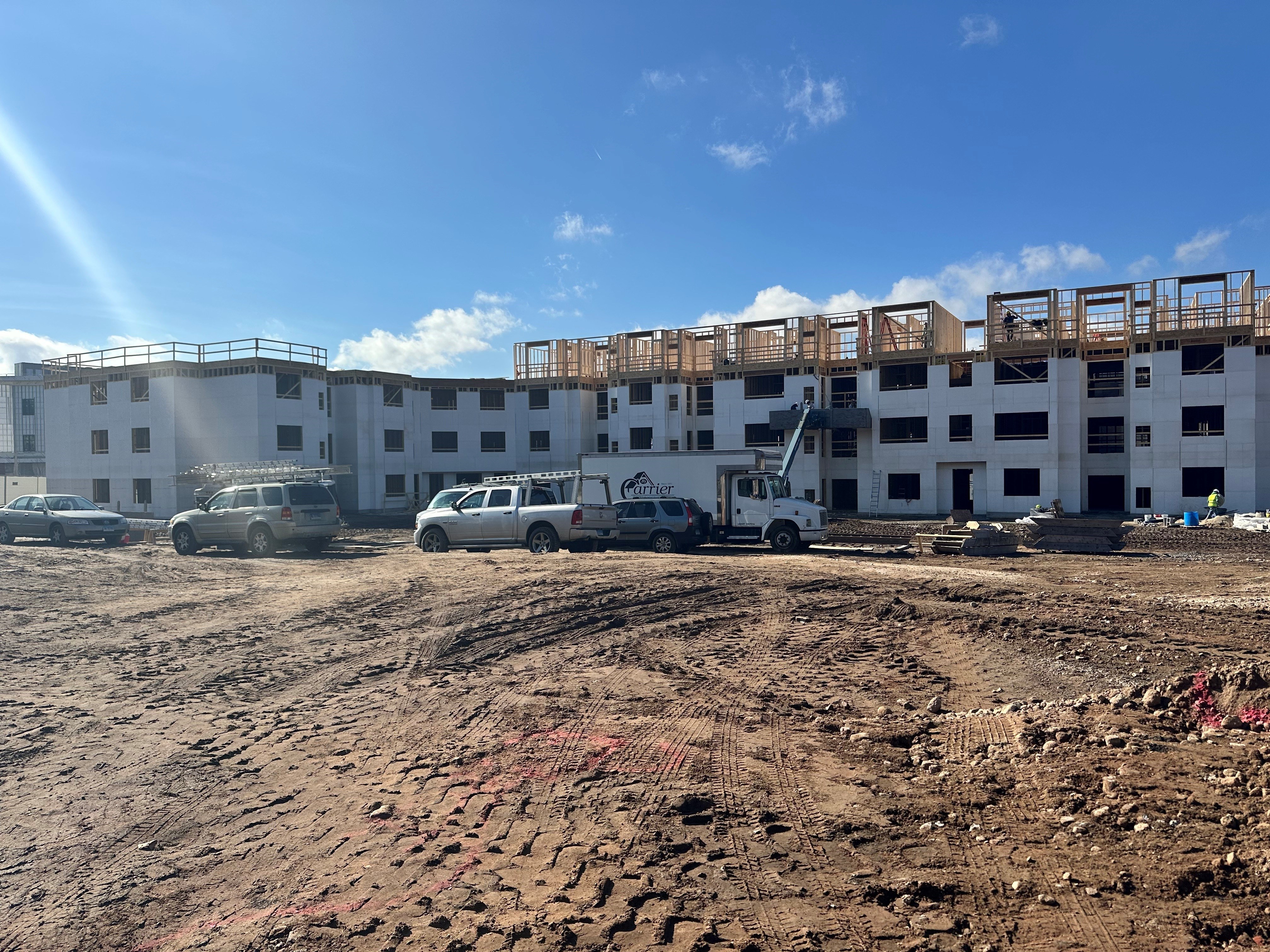 work trucks parked on a field of dirt with tire marks with a white multifamily building under construction